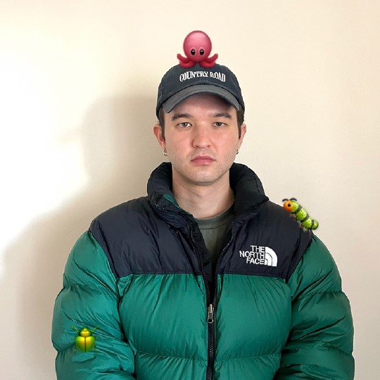 Photo of Paul Rhodes wearing a green and black puffy puffer jacket, he wears a black cap and ontop is a marrone octopus emoji, on his shoulder is a green caterpillar emoji and  on his arm is a green iridescent Christmas beetle